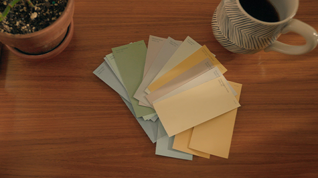 Person holding paint chips up to bathroom wall with sink, mirror and light fixture in background.