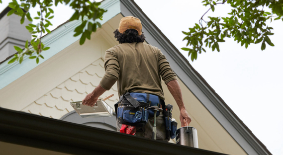 Painter holding paint can, tray and brush looks up to gable roof with creamy neutral scallop siding and gray trim.