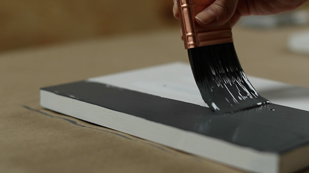 Hand holds a wet brush with dark gray paint and paints a white cabinet door set flat on a table. 